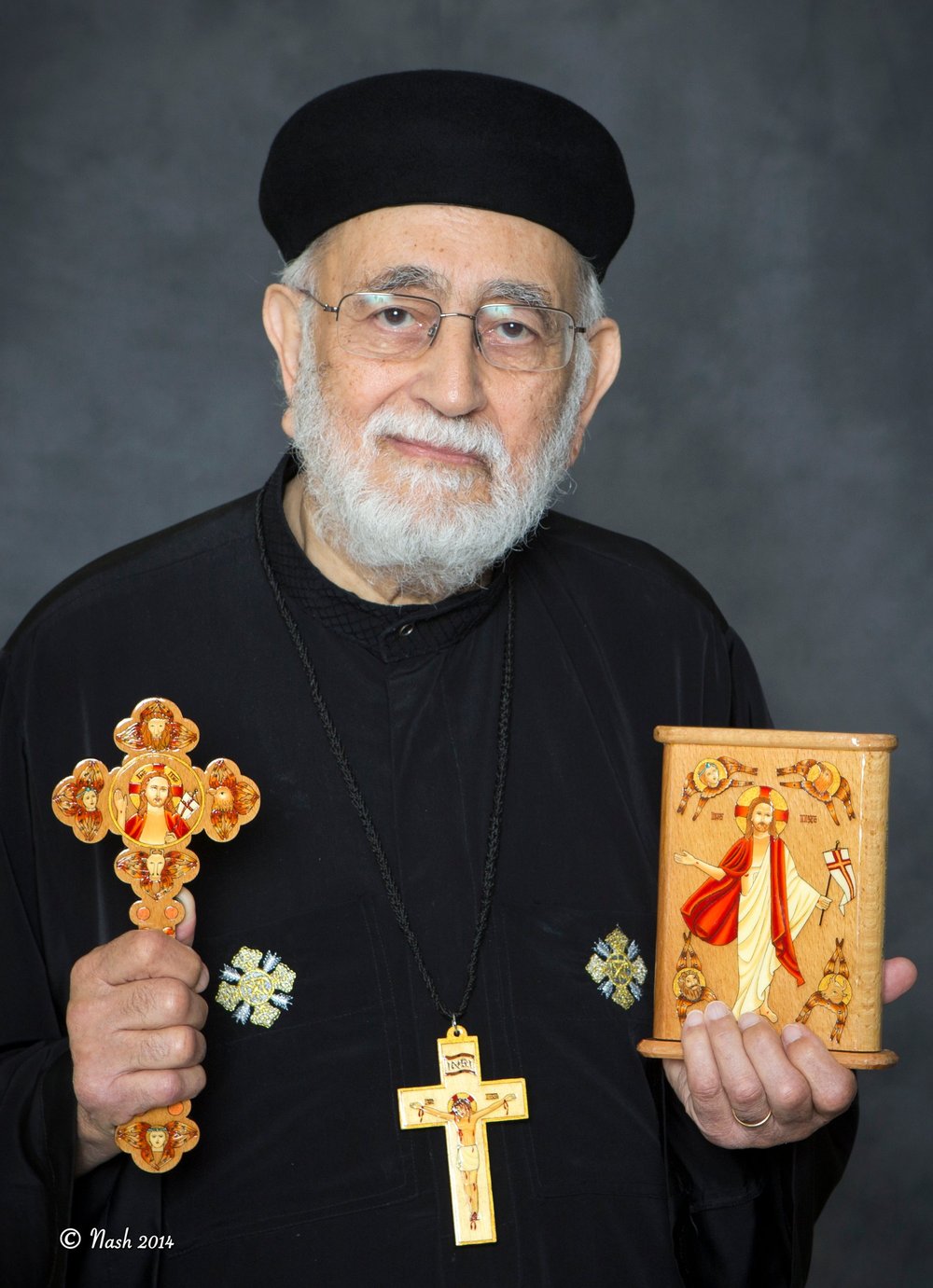 Hegumen Father Mikhail Ibrahim in his black cassock and cap, holding a wooden hand cross in one hand and a small wooden icon of the Resurrection in the other